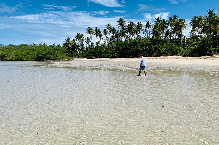 Homem caminhando nas águas transparentes da Praia do Bainema cercada por coqueiros