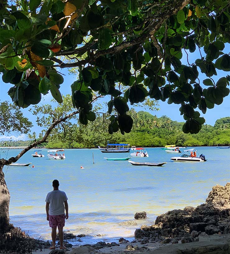 Roteiro em Boipeba: Pessoa observa o mar Moreré com barcos embaixo de árvore e perto de pedras (Foto: Esse Mundo é Nosso)