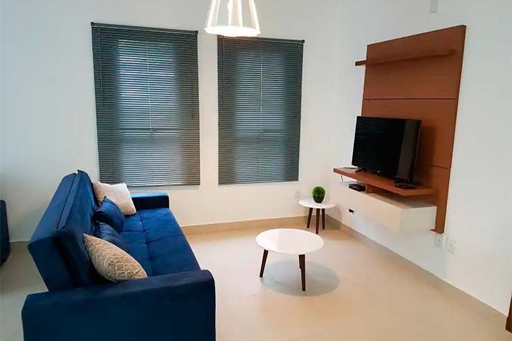 Sala de estar da casa com TV e sofá azul, além de mesa de centro (Foto: Divulgação)