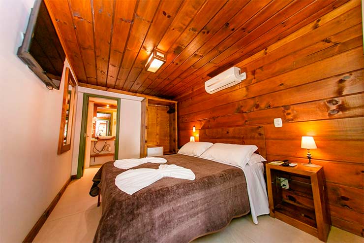 Quarto da Pousada Charm Canela com cama de casal, parede e teto de madeira e TV (Foto: Divulgação)