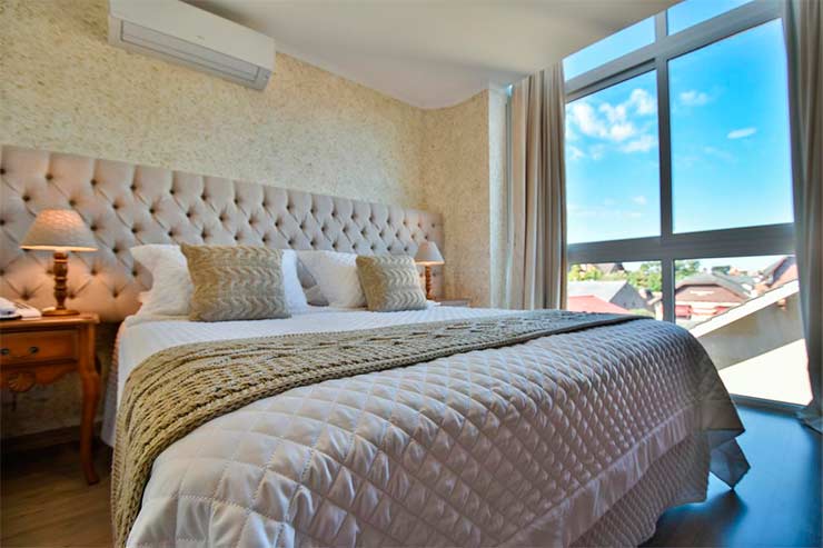 Acomodação do Micheline Hotel Tricot com cama de casal e grande janela com céu azul (Foto: Divulgação)