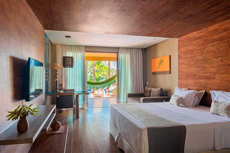 Acomodação do Carmel Charme Resort com cama de casal, TV e varanda em tons amadeirados