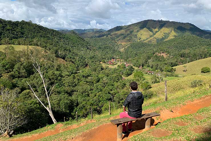 Homem sentado no mirante do Vale do Alcantilado com vista para as montanhas em dia de sol e nuvens