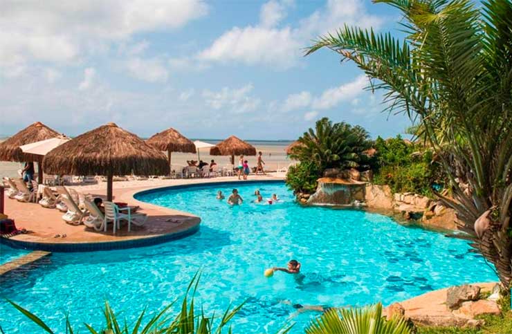 Pessoas brincam na piscina do Vila Galé  Resort do Cabo e outras descansam em espreguiçadeiras em dia de céu azul com algumas nuvens