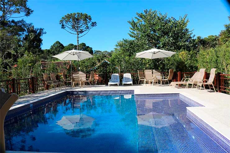 Piscina da pousada Jardim da Luz com cadeiras e guarda-sol em meio a árvores em dia de céu azul