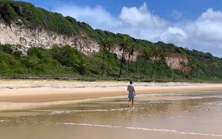 Homem caminha pelo mar da Praia do Madeiro em dia de céu azul