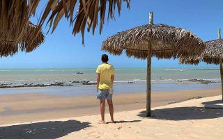 Homem observa o mar esverdeado do alto ao lado de guarda-sol na praia de Monte Alegre, no RN