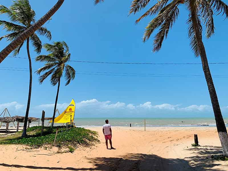 Homem em pé ao lado de árvores com mar à frente em dia de céu azul em Perobas, perto de São Miguel do Gostoso