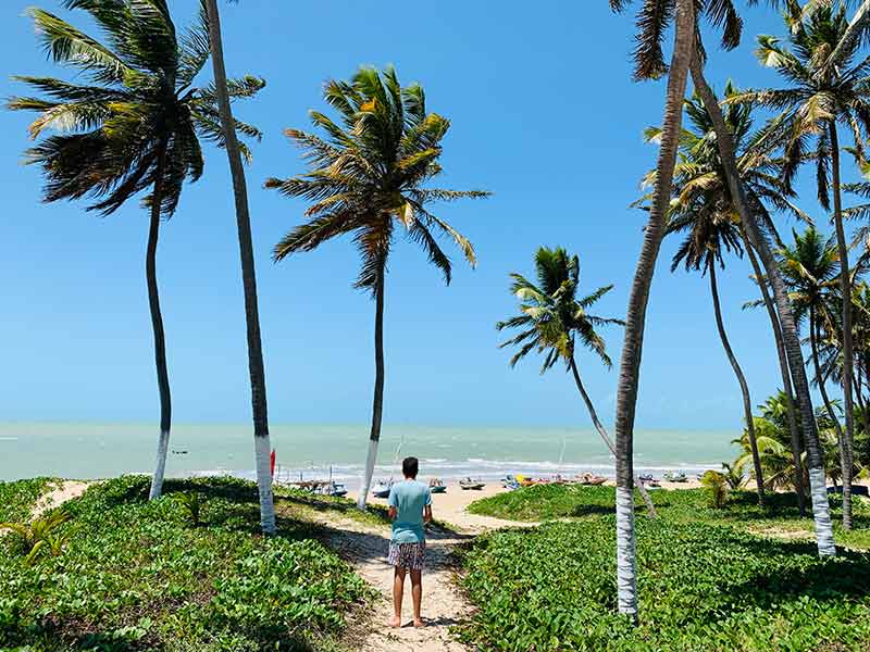 Homem passa por caminho de árvores rumo ao mar esverdeado da praia de Zumbi, perto de São Miguel do Gostoso