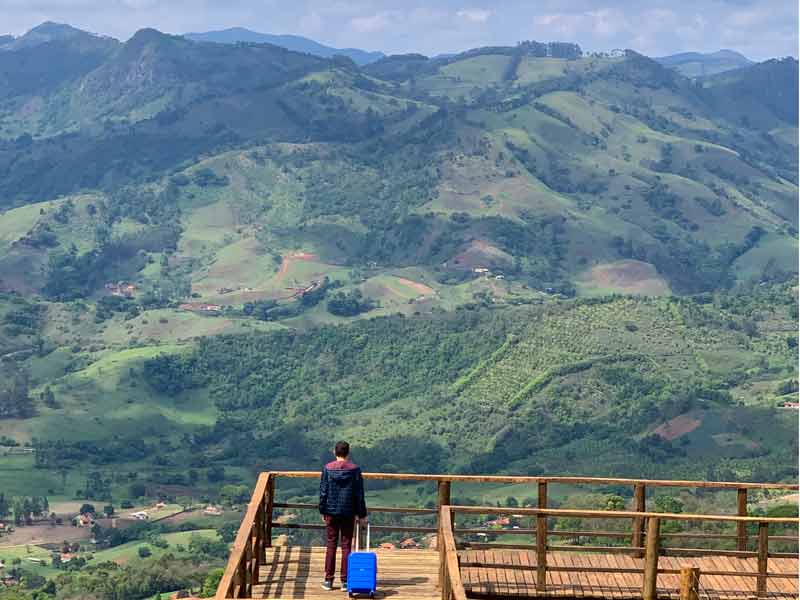 Homem segura mala azul em mirante com vista das montanhas