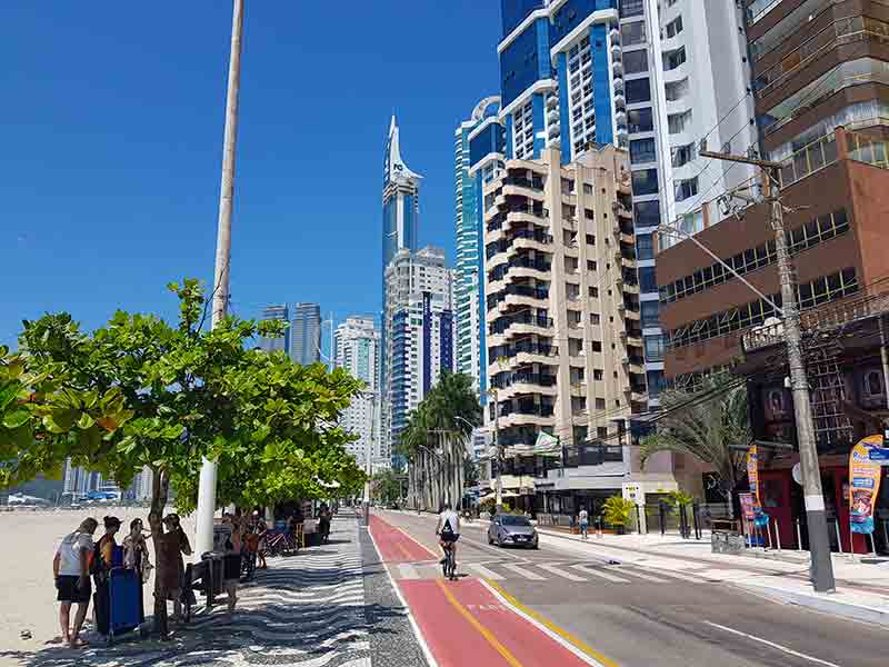 Pessoas curtem dia de sol enquanto carro e bicicleta passeiam pela orla de Balneário Camboriú