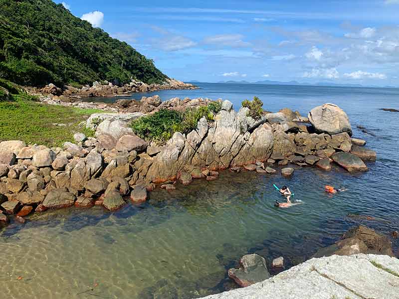 Pessoas se divertem no mar na Ilha do Macuco, em Bombinhas, SC, em dia de céu azul