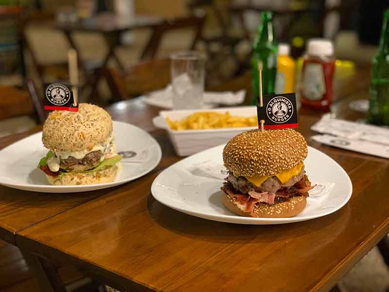 Sanduíche Suíço e Costela Burger com batata frita ao fundo do O Alquimista em Bombinhas, SC