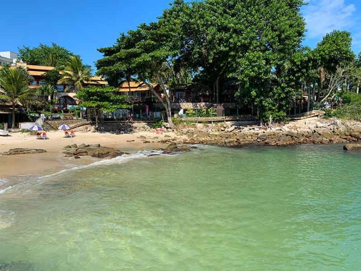 Mar esverdeado vazio com árvores à frente em dia de céu azul na Prainha, em Bombinhas