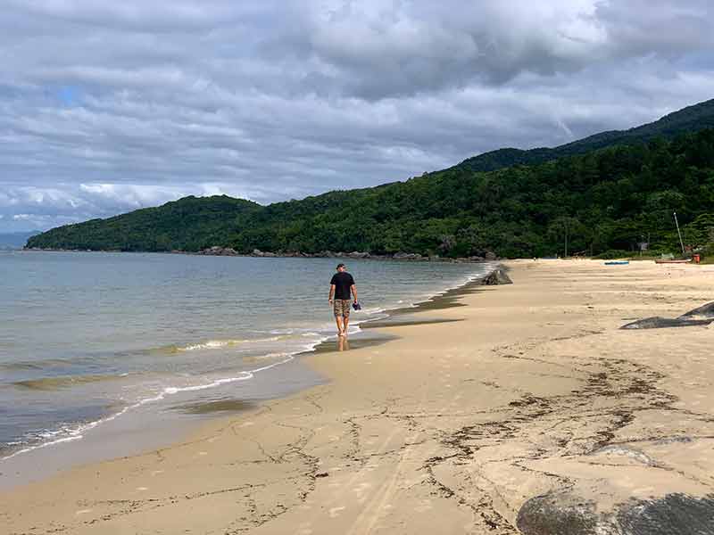 Homem caminha sozinho pela praia de Zimbros, em Bombinhas, SC, em dia nublado