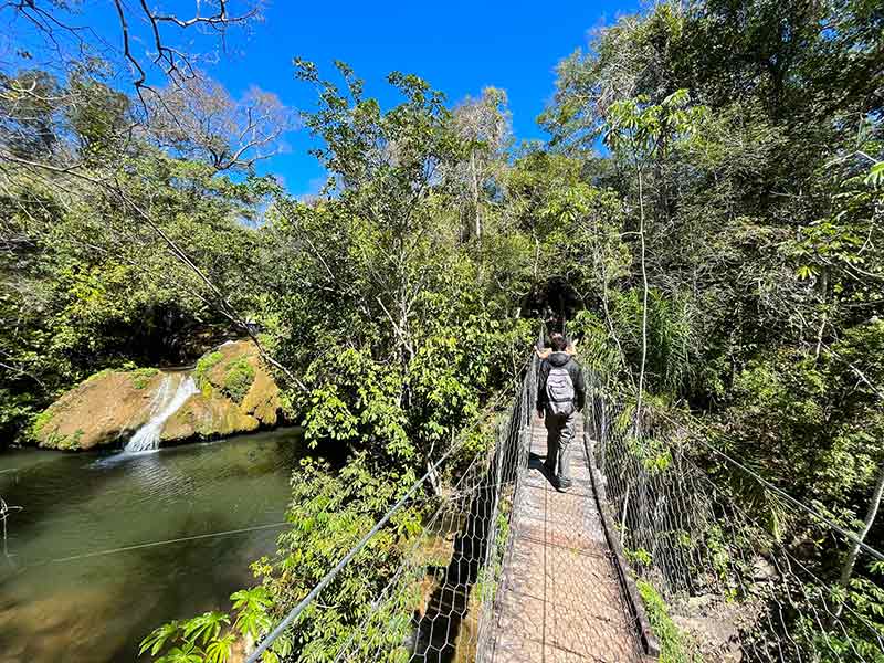 Homem na Ceita Corê, em Bonito, MS, passa por ponte suspensa com mochila, casaco e calça com cachoeira ao lado em dia de céu azul