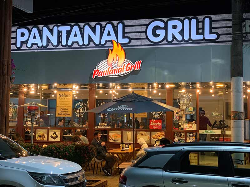 Pessoas sentadas na parte externa do Pantanal Grill, em Bonito, com carros parados na porta