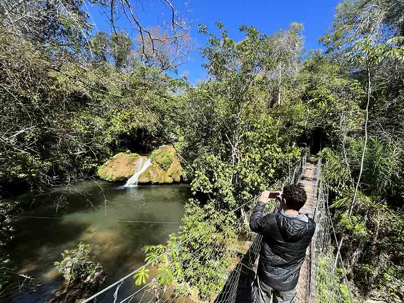 Quando ir para Bonito: Homem atravessa ponte suspensa com cachoeira na Ceita Coré