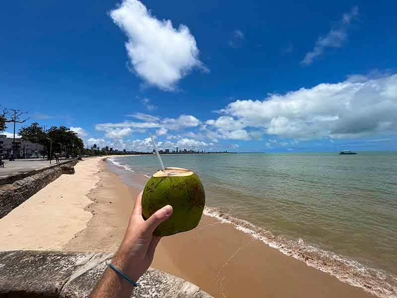Homem segura água de coco em praia em João Pessoa em dia de céu azul com poucas nuvens
