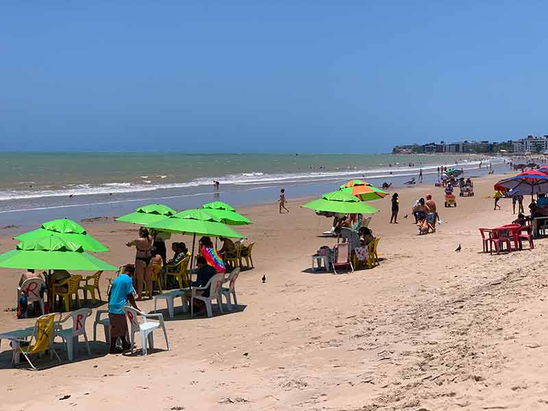 Pessoas sentadas em guarda-sóis verdes no Bessa, uma das praias em João Pessoa, em dia de céu azul