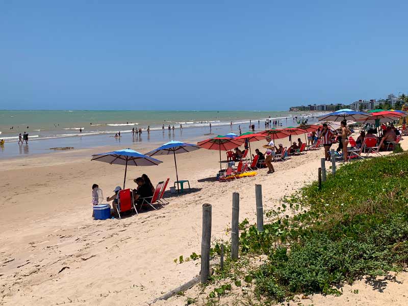 Pessoas com guarda-sóis em dia de céu azul nas areias da Praia do Bessa