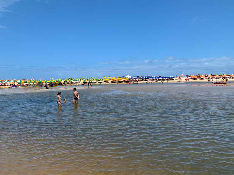 Pessoas se divertem no rio com guarda-sóis ao fundo na Praia Bela