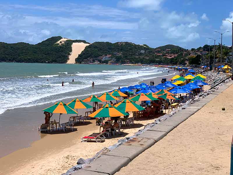 Guarda-sóis coloridos na areia, pessoas no mar e Morro do Careca no canto em Ponta Negra, uma das praias de Natal