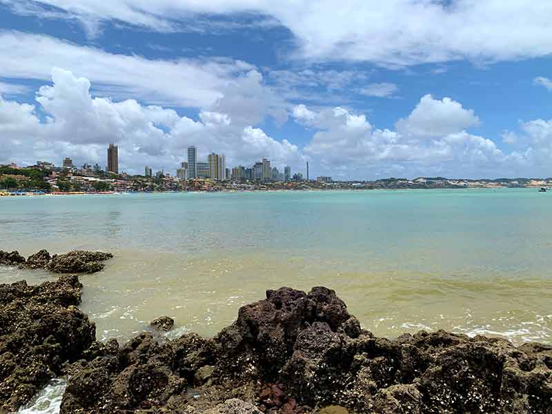 Prédios ao fundo, mar e pedras em dia com algumas nuvens em Ponta Negra