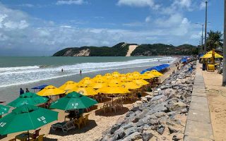 Guarda-sóis verdes e amarelos em Ponta Negra, uma das praias de Natal, com Morro do Careca ao fundo e pessoas no mar