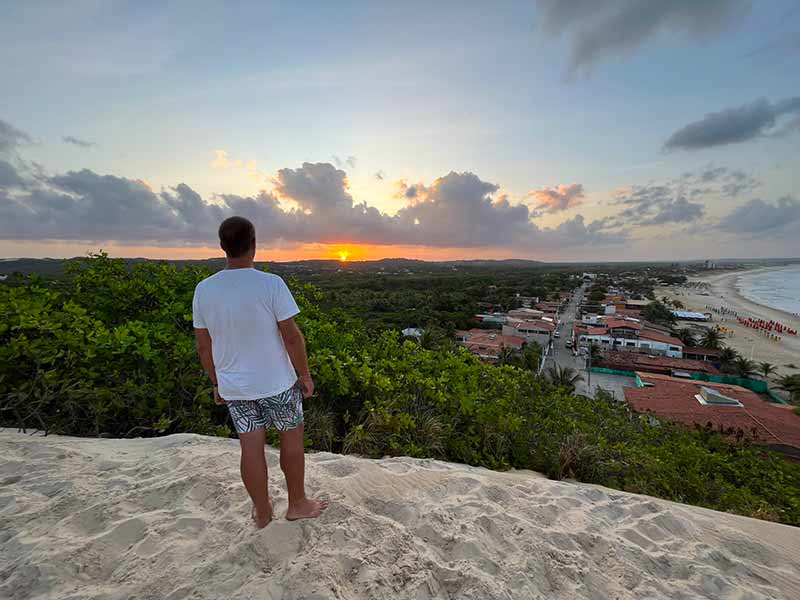 Homem em pé nas Dunas de Genipabu observa o pôr do sol