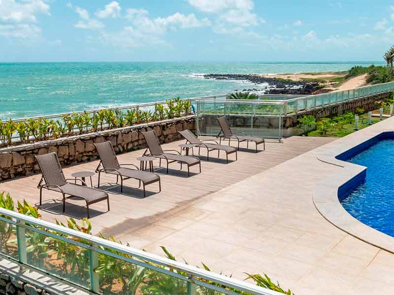 Área da piscina do Senac Barreira Roxa, dica de hotel em Natal, com espreguiçadeiras vazias e mar ao fundo
