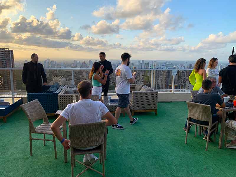 Homem sentado em mesa e pessoas andando ao ar livre na parte externa do Skybar, em João Pessoa