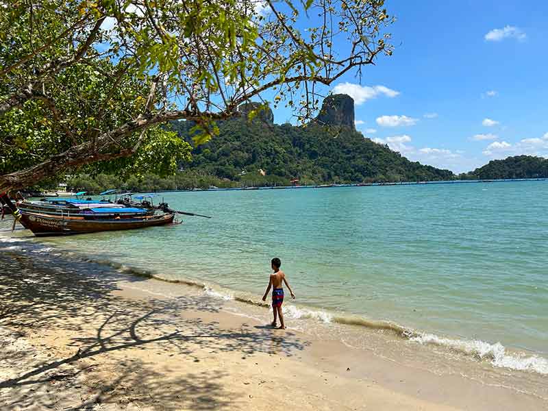 Criança anda na areia da East Railay perto dos barcos e com pedras ao fundo