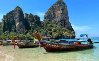 Barcos parados no mar de West Railay, em Railay Beach, na Tailândia, com pedras ao fundo e céu azul