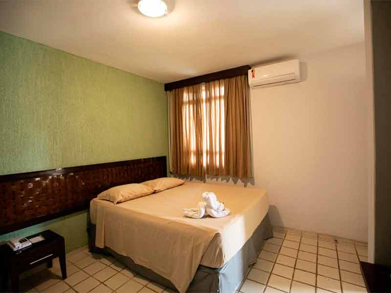Quarto simples do Eco Chalés, dica de onde ficar em Rio Quente, com cama de casal, janela com cortina escura, parede verde e ar-condicionado