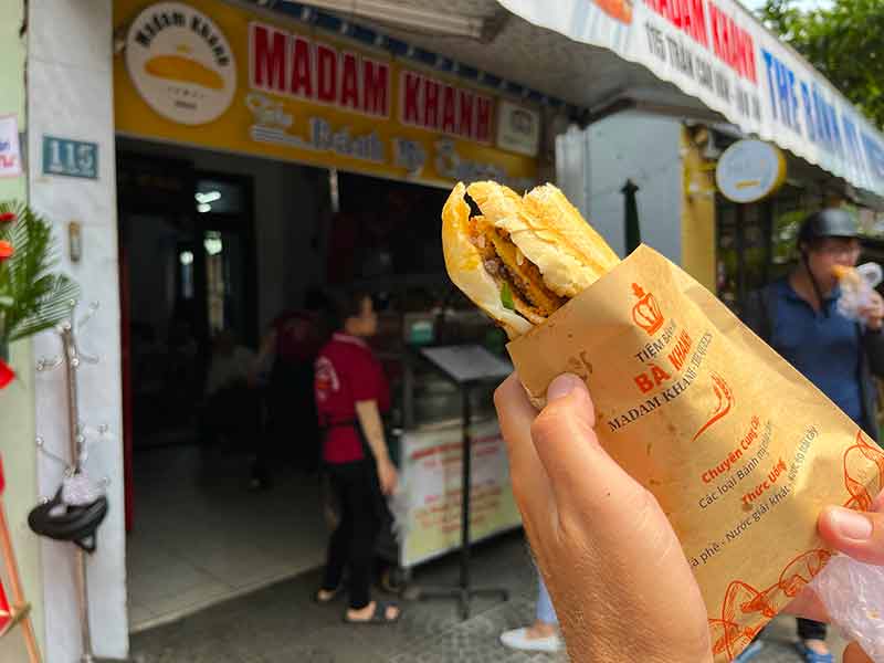 Homem segura báhn mi, baguete vietnamita, em frente à entrada de lanchoete