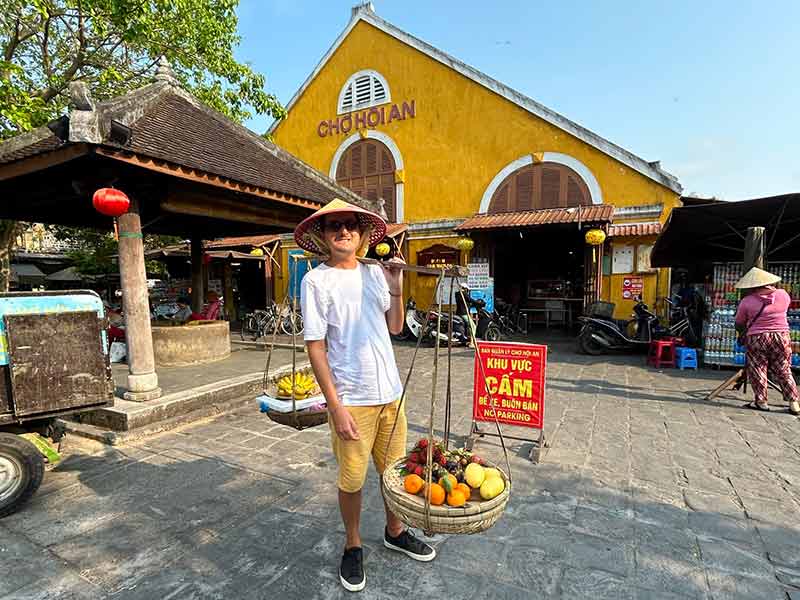 Homem usa chapéu do Vietnã e tira foto segurando cestas de frutas no ombro, dica de o que fazer em Hoi An