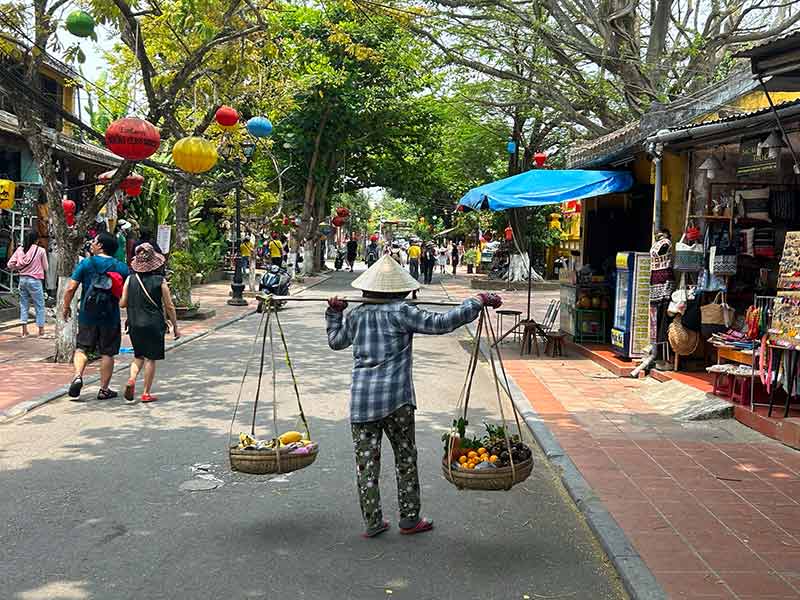 Mulher segura dois cestos de frutas nos ombros em meio a rua de cidade no Vietnã