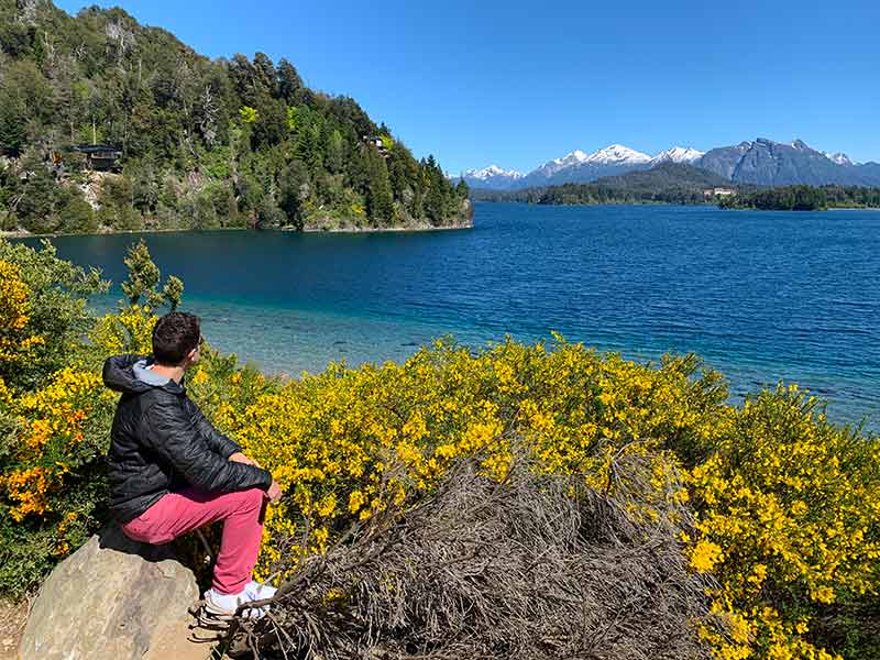 Homem sentado em pedra observa lago e montanhas no Circuito Chico, em Bariloche, na primavera