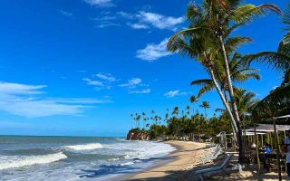 Praia da Barra do Cahy, na Bahia, com coqueiros, grande faixa de areia e mar com ondas