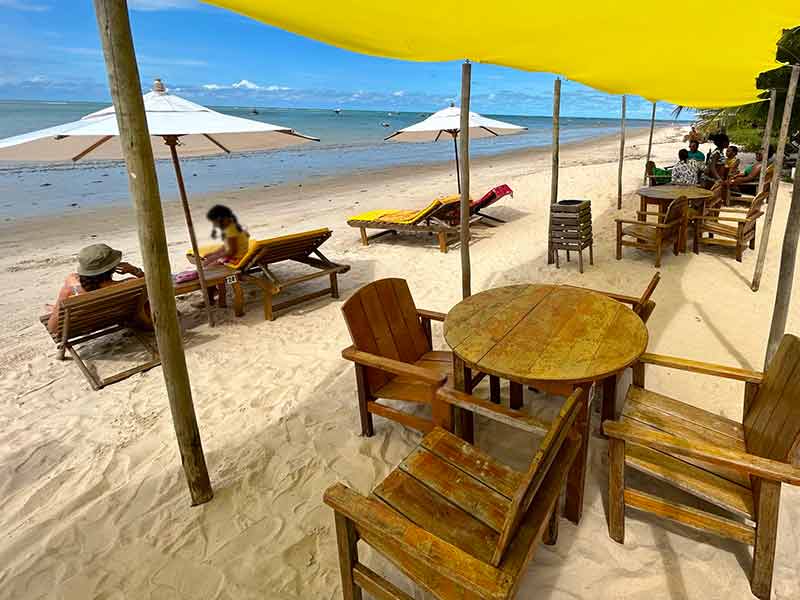 Área externa do Binotto's, em Cumuruxatiba, na areia da praia central com tendas amarelas e mesas