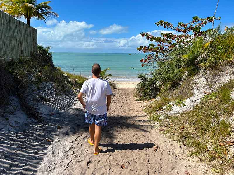 Homem caminha pela areia no sentido da praia de Corumbau, na Bahia, em dia de céu azul