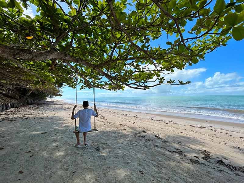 Homem em balanço embaixo de árvore na praia de Corumbau, na Bahia, em dia de céu azul com poucas nuvens