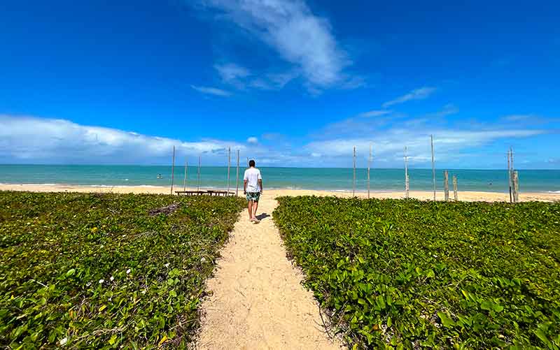 Homem caminha em areia entre vegetações rumo à praia da Ponta do Corumbau em dia de céu azul