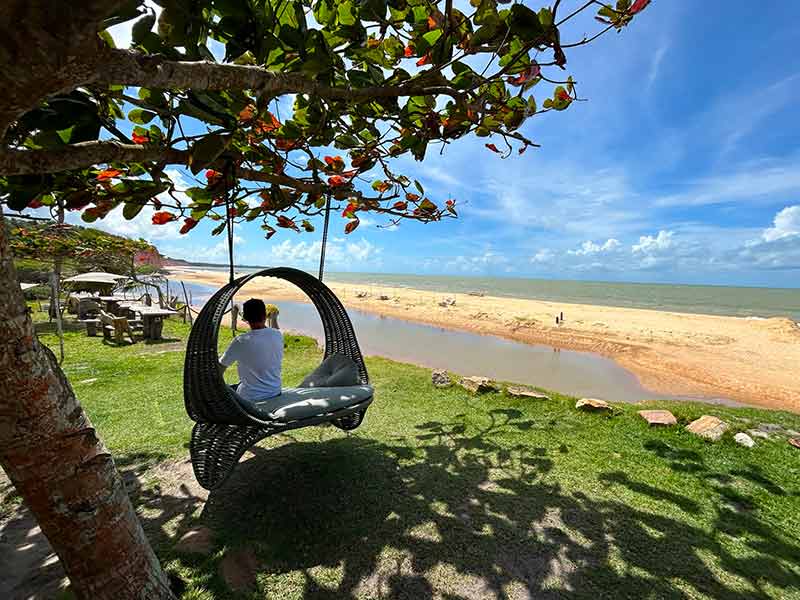 Homem sentado em balanço em Japara Grande, em Cumuru, observa rio e mar em dia de céu azul