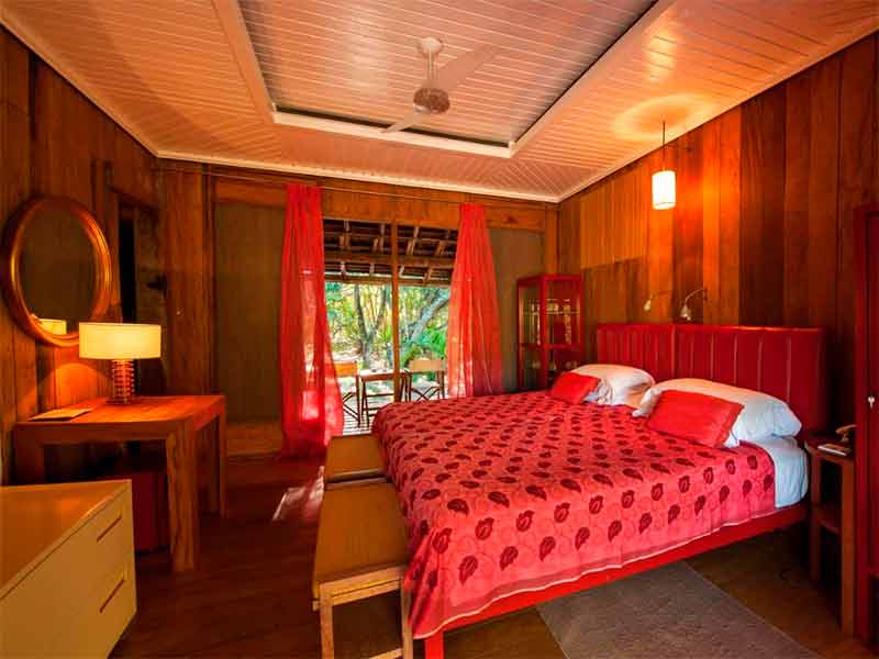 Quarto de madeira da Vila Naiá, dica entre as pousadas em Corumbau, com cama de casal com roupa vermelha