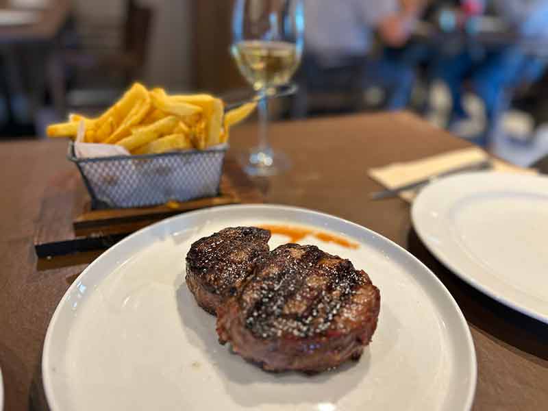 Ojo de bife com batatas fritas ao fundo e taça de vinho branco do Mi Viejo