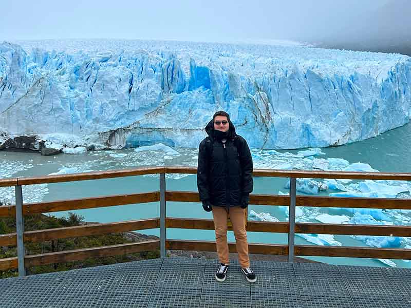 Homem posa em passarela em frente ao Perito Moreno, que tem tons brancos e azuis