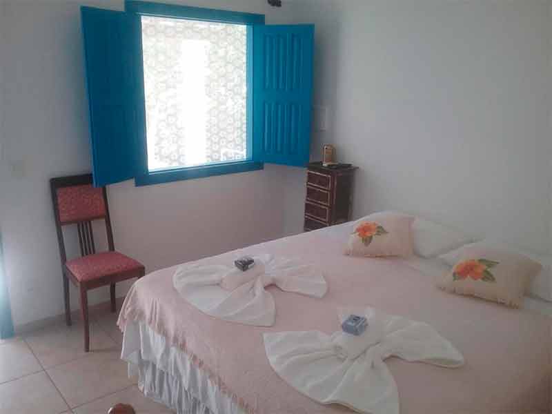 Quarto simplesa da PraiAmar com cama de casal, janela azul e cadeira encostada na parede