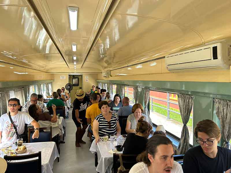 Vagão restaurante com pessoas sentadas em mesas para quatro em trem de Guararema
